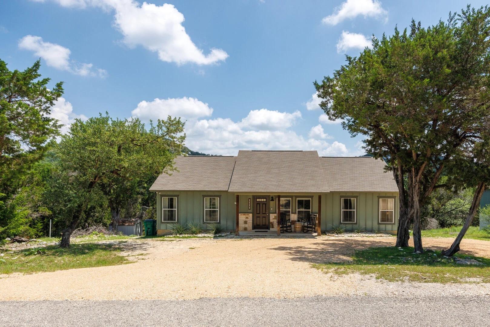 Front of the green board-and-batten cottage with a covered porch, framed by oak and cedar trees.