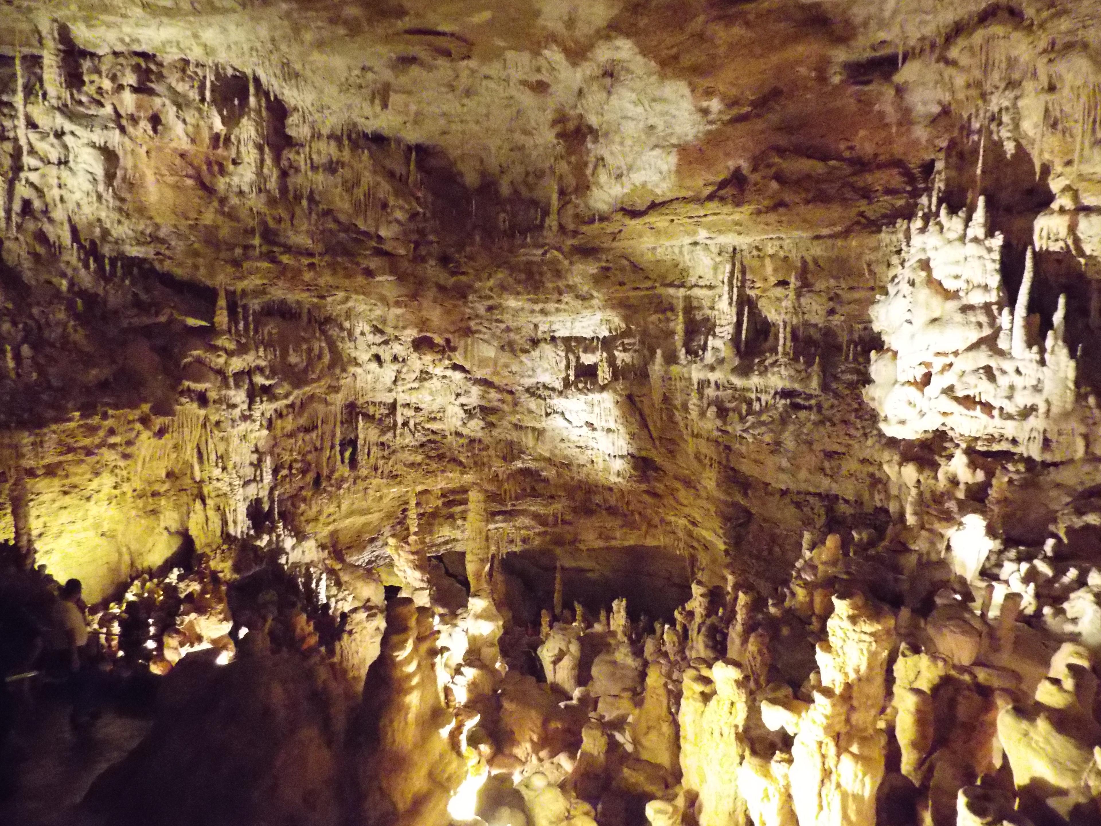 A vast underground cavern chamber lit to show stalactites and stalagmites, with tiny visitors for scale